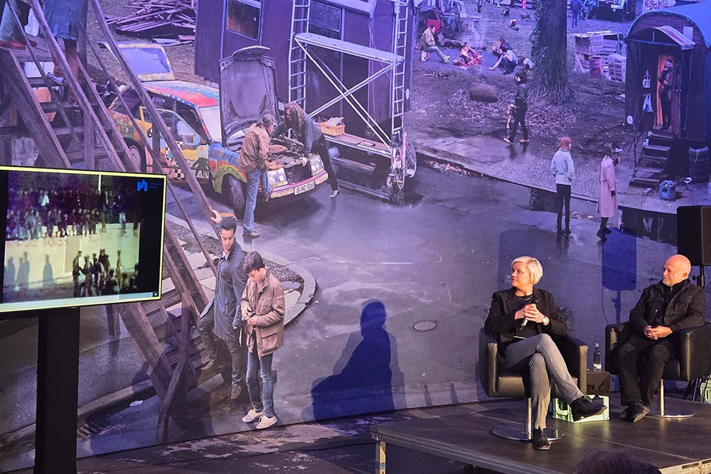 Links auf der Bühne ist Dr. Susanne Muhle, Projektleiterin am Standort Checkpoint Charlie, zu sehen, wie sie mit Yadegar Asisi, rechts im Bild, ein Gespräch anlässlich des 9. Novembers 2024 führt. Hinter der kleinen Bühne ist das riesige Panorama zu erkennen, das der Künstler Asisi entworfen hat